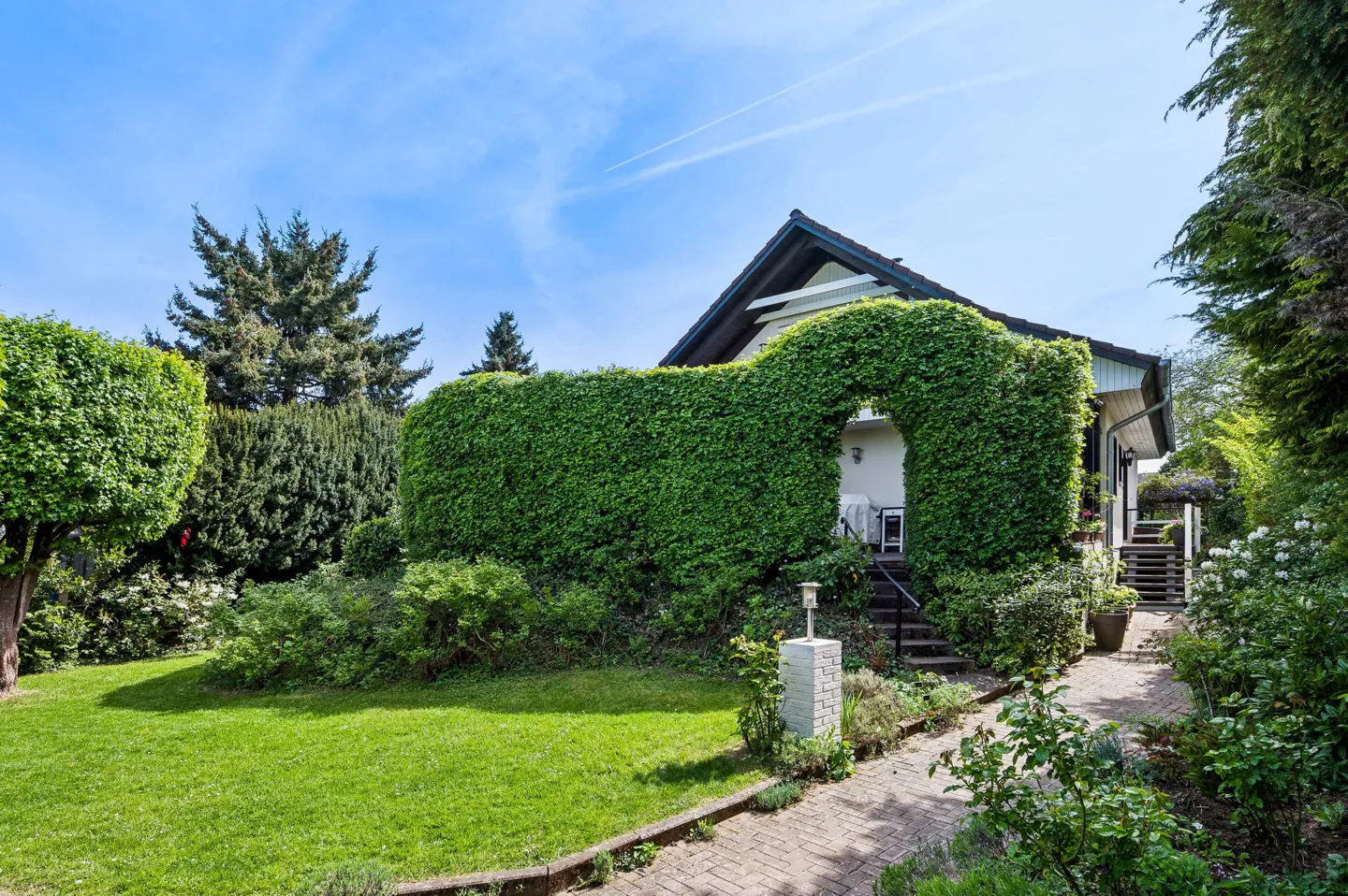 A house with a green hedge archway over the entrance, a brick path, and a green lawn under a blue sky.