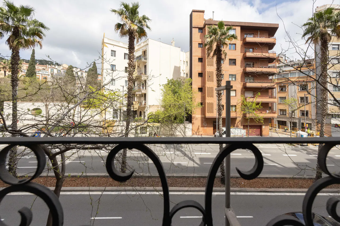 View from a balcony with black wrought iron railing overlooking a city street with palm trees and buildings.