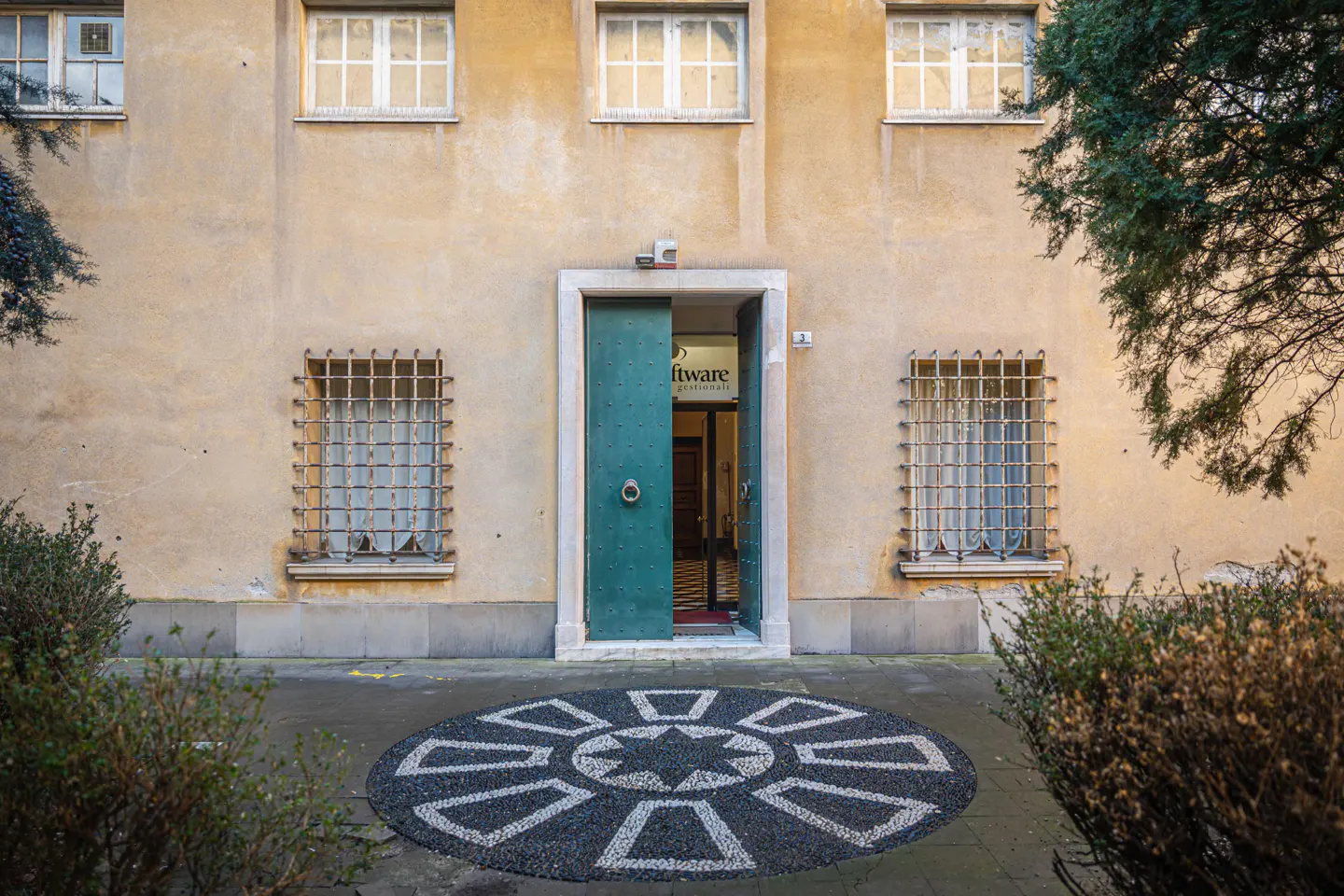 Exterior of a yellow building with a teal door ajar, revealing an office. Windows have metal bars. A stone mosaic is on the ground.