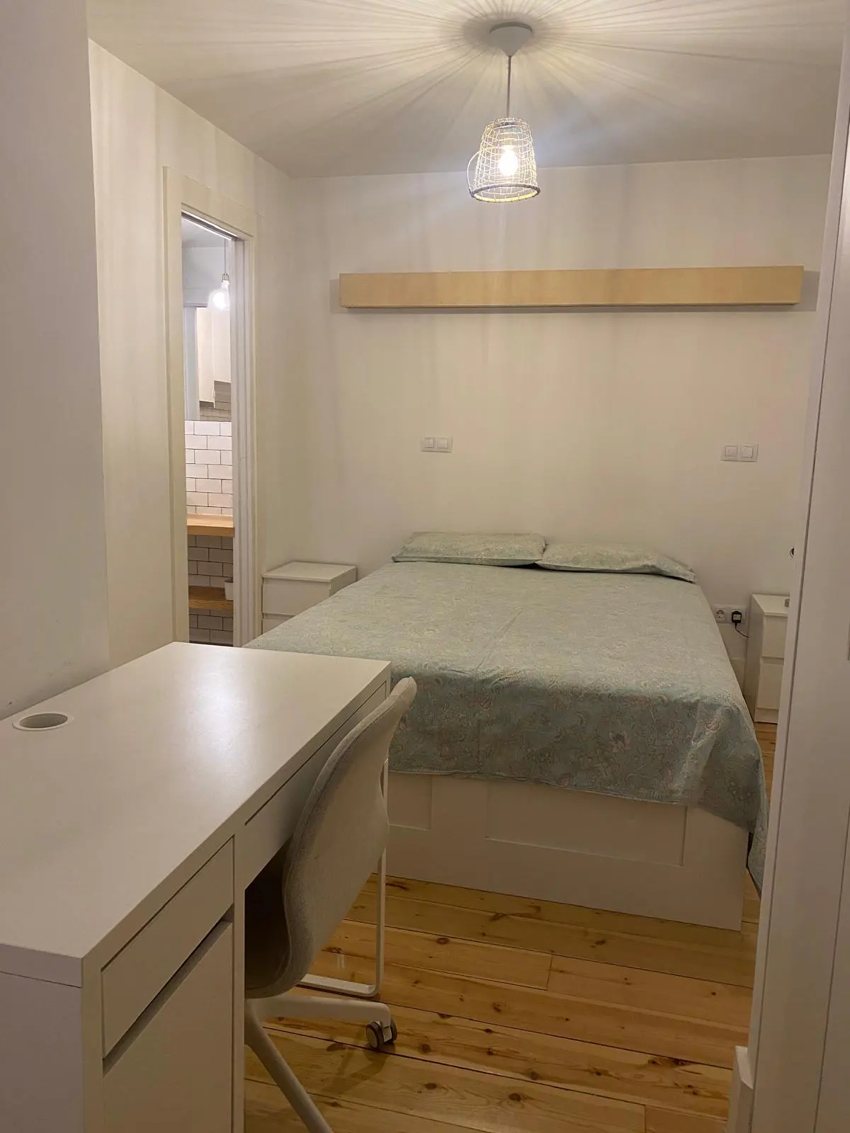 Bedroom with a double bed, white desk and chair, and wooden floors. A doorway leads to a kitchen with white brick tiles.