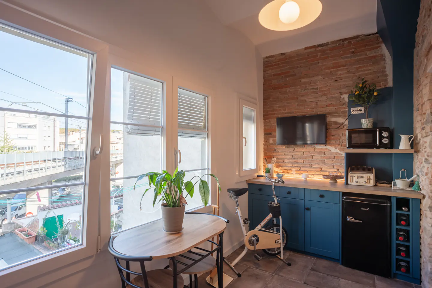 Cozy apartment kitchen with brick wall, blue cabinets, and a small dining table by a large window. An exercise bike stands nearby.