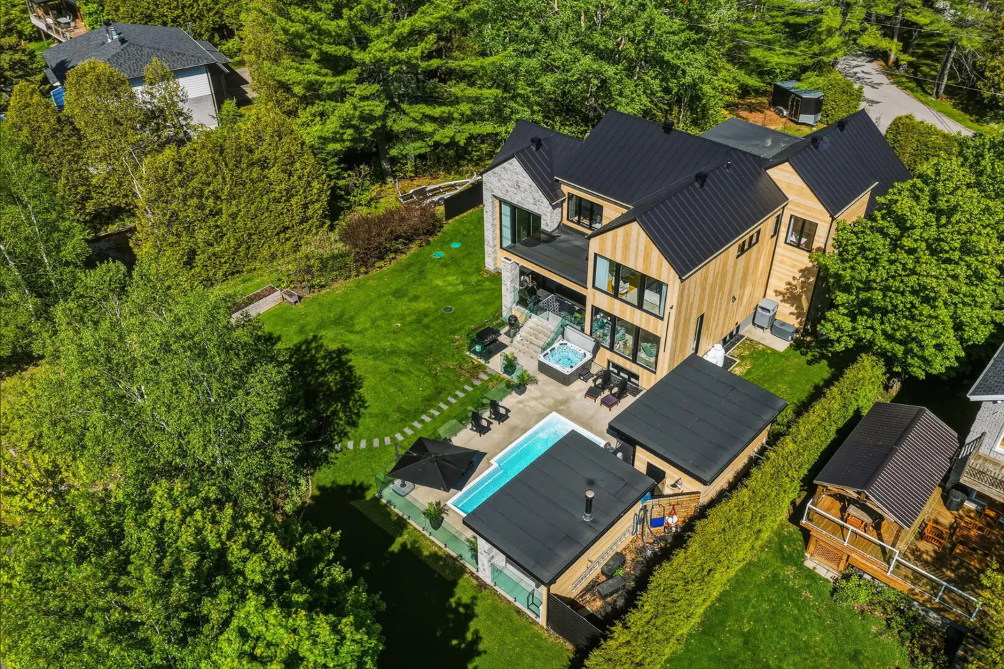 Aerial view of a modern wood-sided home with a black metal roof, swimming pool, hot tub, and lush green backyard.
