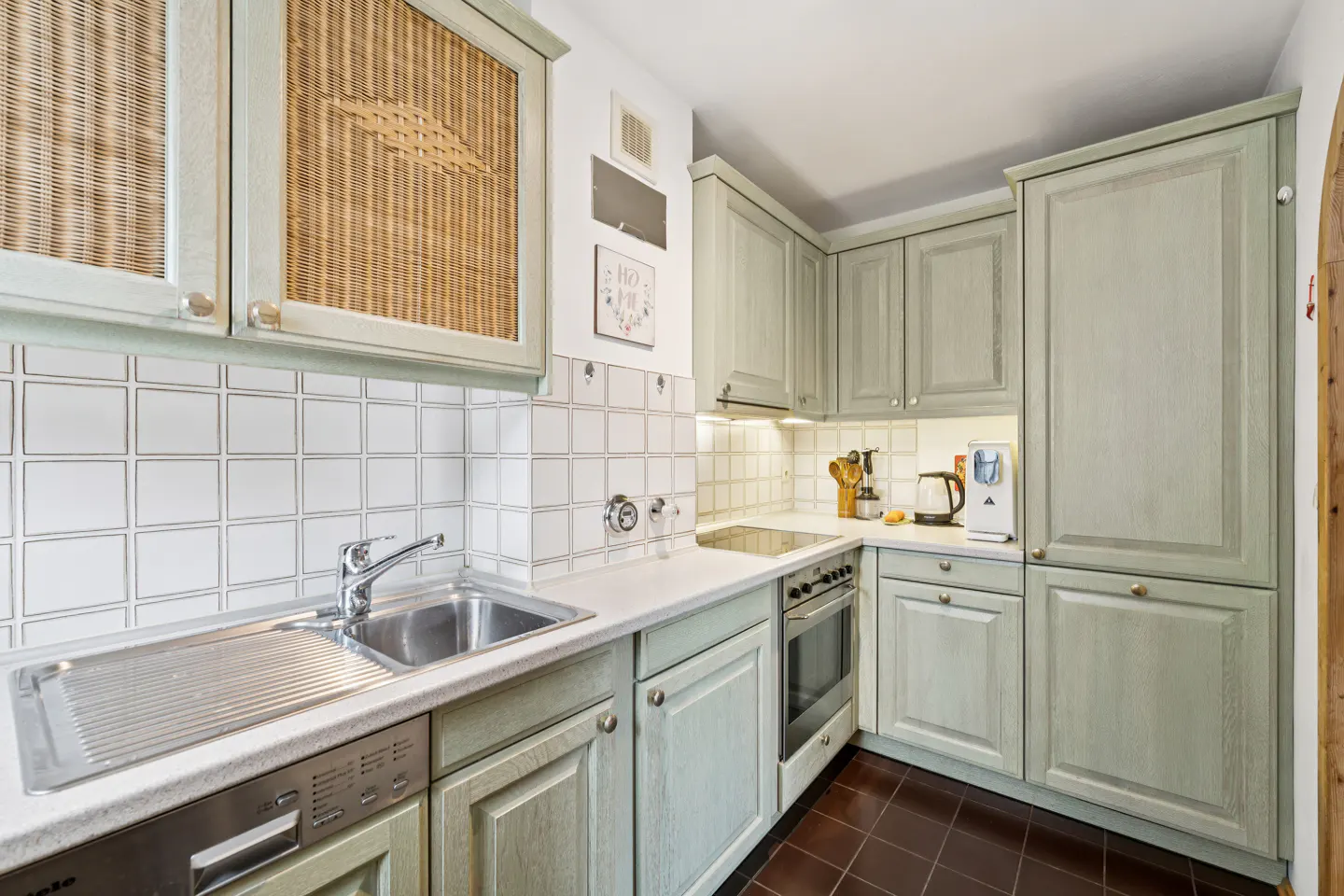 A kitchen with light green cabinets, white countertops, and a stainless steel sink. The upper cabinets have woven rattan inserts. Brown tile floor.