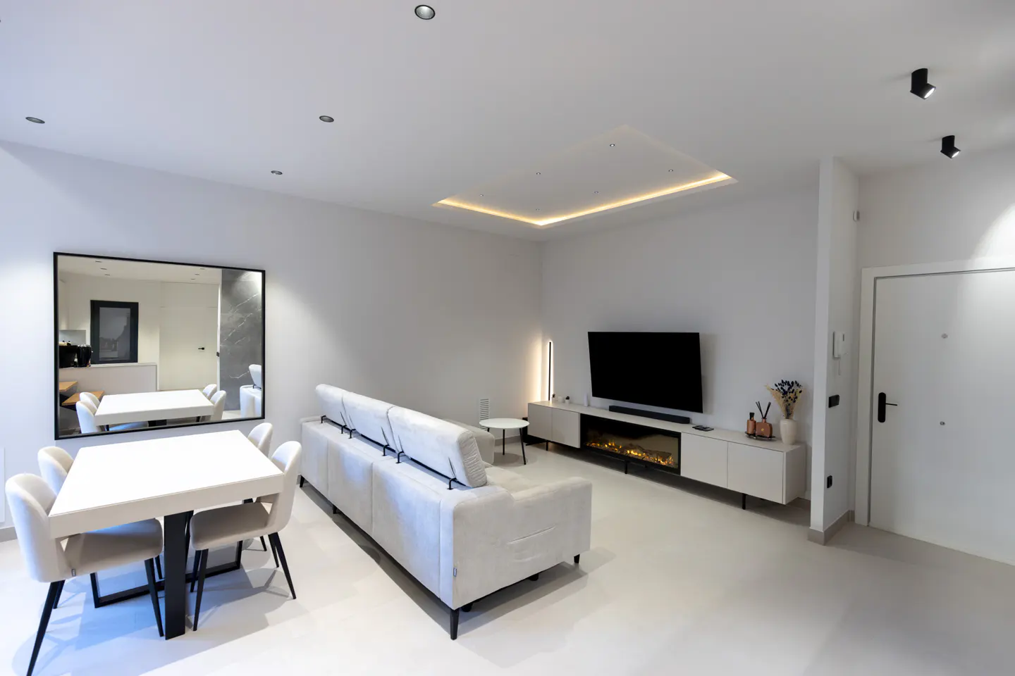 Bright, modern living room with white walls, a sofa, a TV, and a dining table with chairs. A large mirror reflects the dining area.