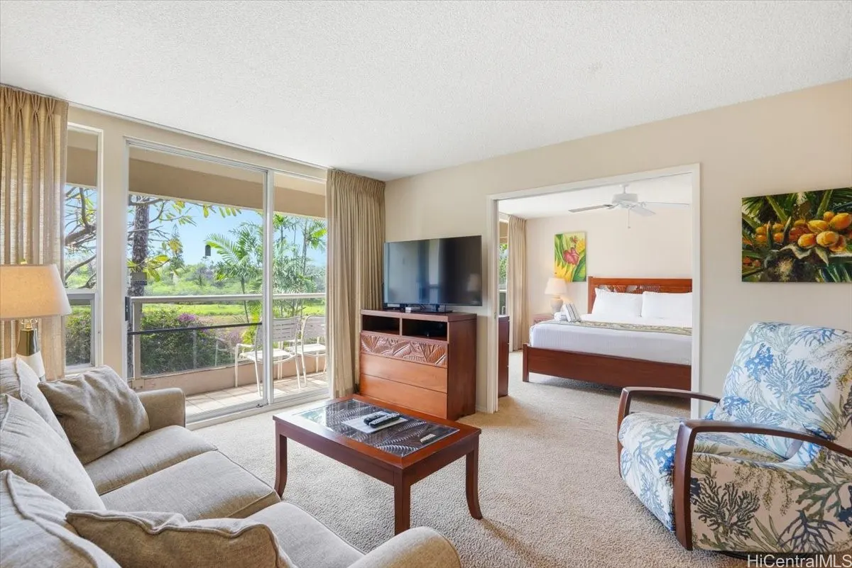 Living room with beige sofa, wood table, and TV. Balcony with green view. Bedroom visible through doorway with bed and floral art.