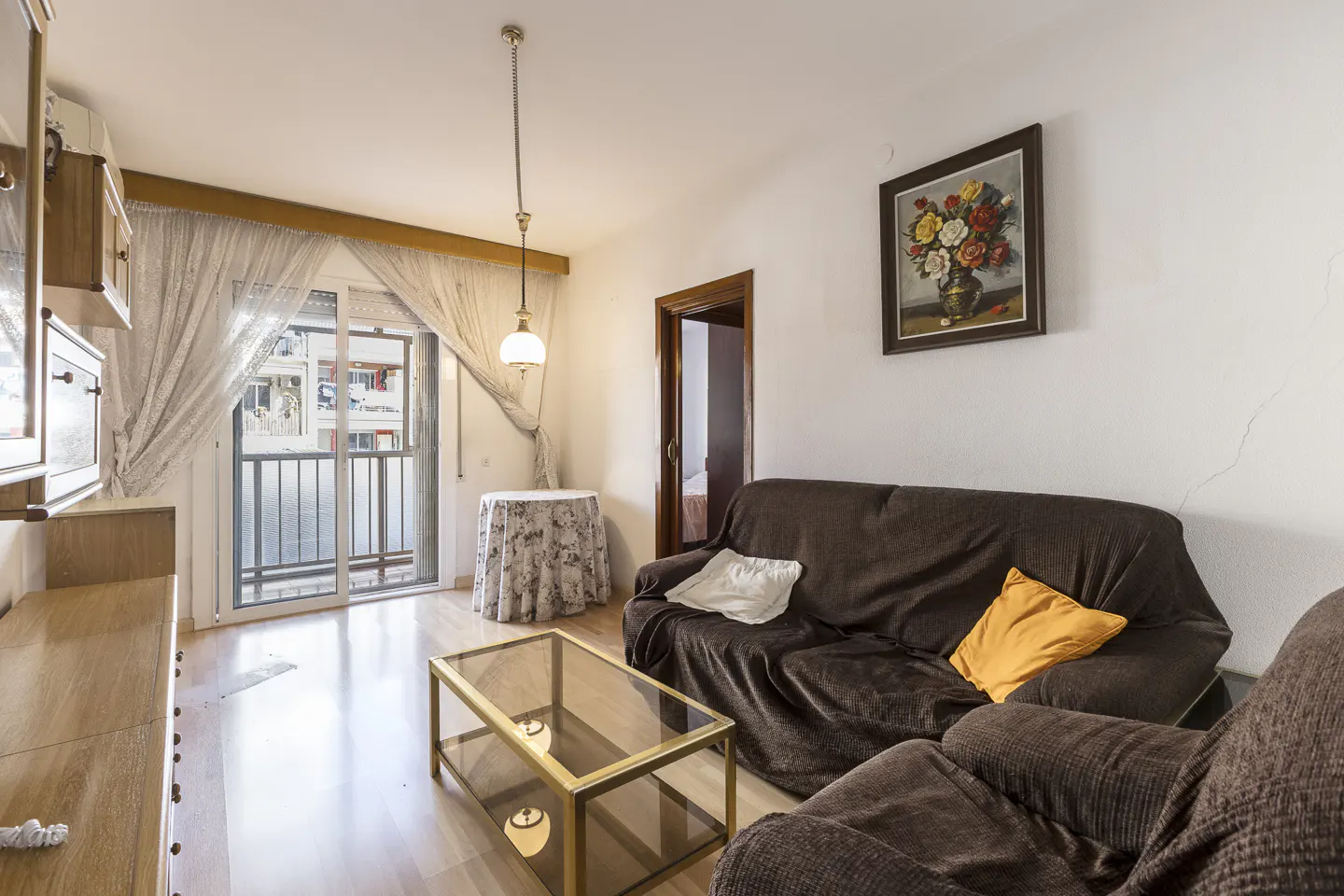 Living room with brown sofa, glass table, and balcony. A flower painting hangs on the white wall. Light wood floors.