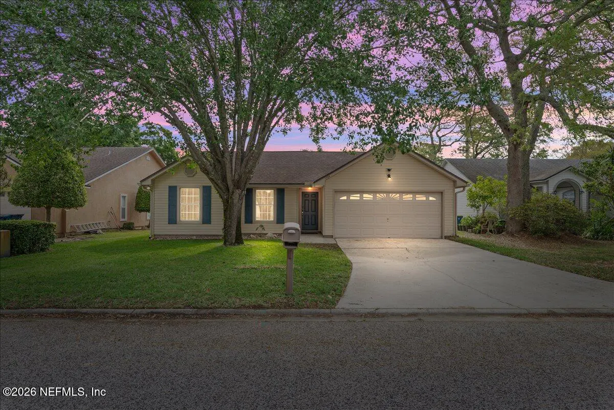 Beige single-story house with blue shutters, a driveway, and a green lawn under a pink and purple sky.