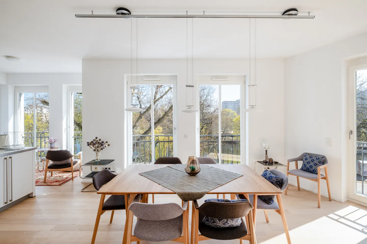 Bright dining room with a light wood table, gray chairs, and pendant lights. Balcony doors offer a view of trees.