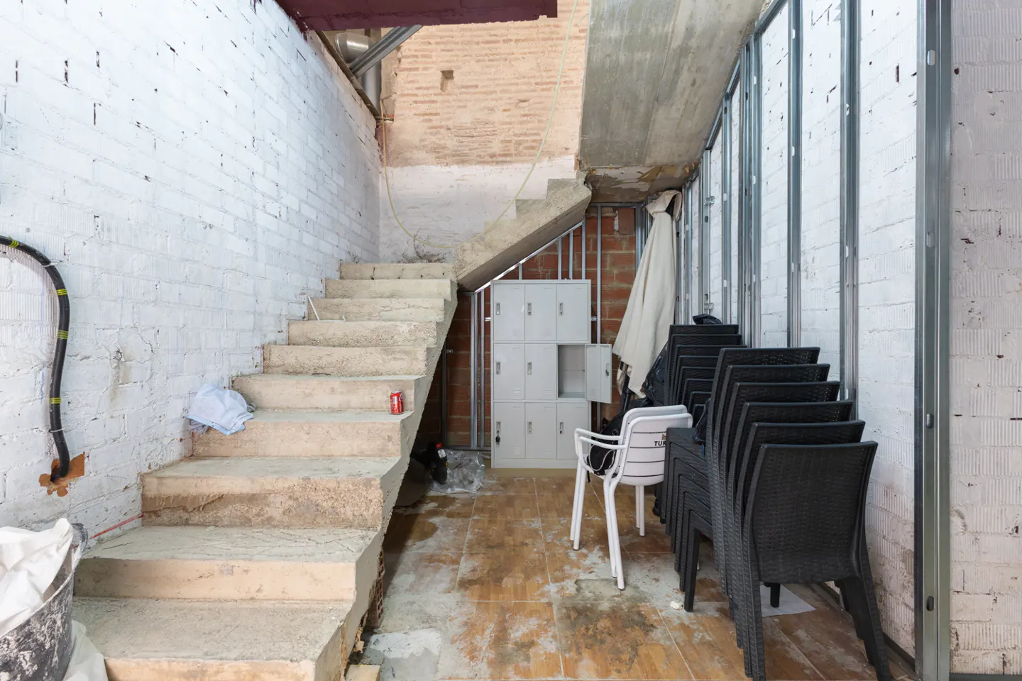 Concrete staircase in a room under renovation with white brick walls, metal framing, stacked black chairs, and lockers.