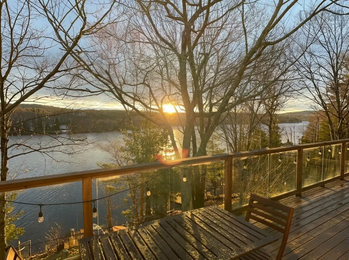 Wooden deck with table and chair overlooks a lake at sunset. String lights hang along the glass railing. Bare trees frame the view.