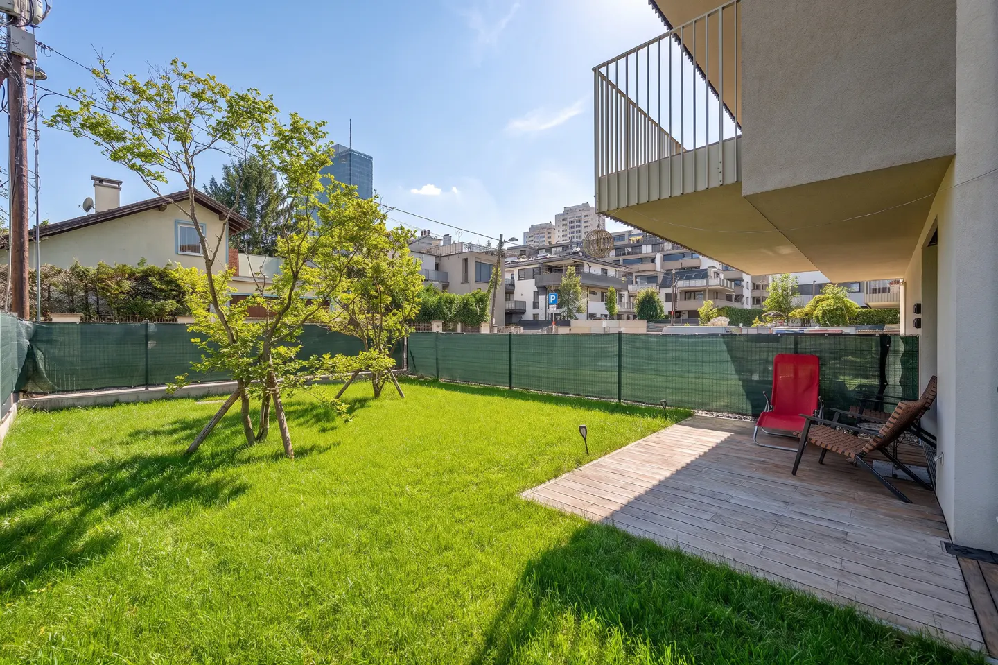 A modern home's backyard features a green lawn, young trees, a wooden deck with chairs, and a green fence.