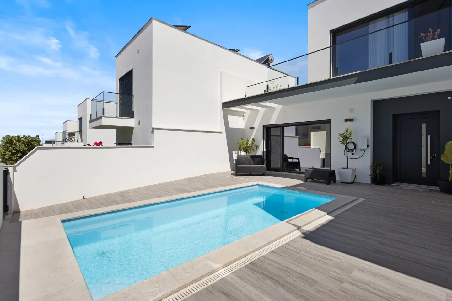 A modern white house with a blue pool and wooden deck under a bright blue sky.