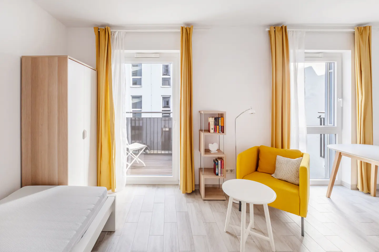 Bright studio apartment with yellow curtains, chair, and white furniture. Balcony view.