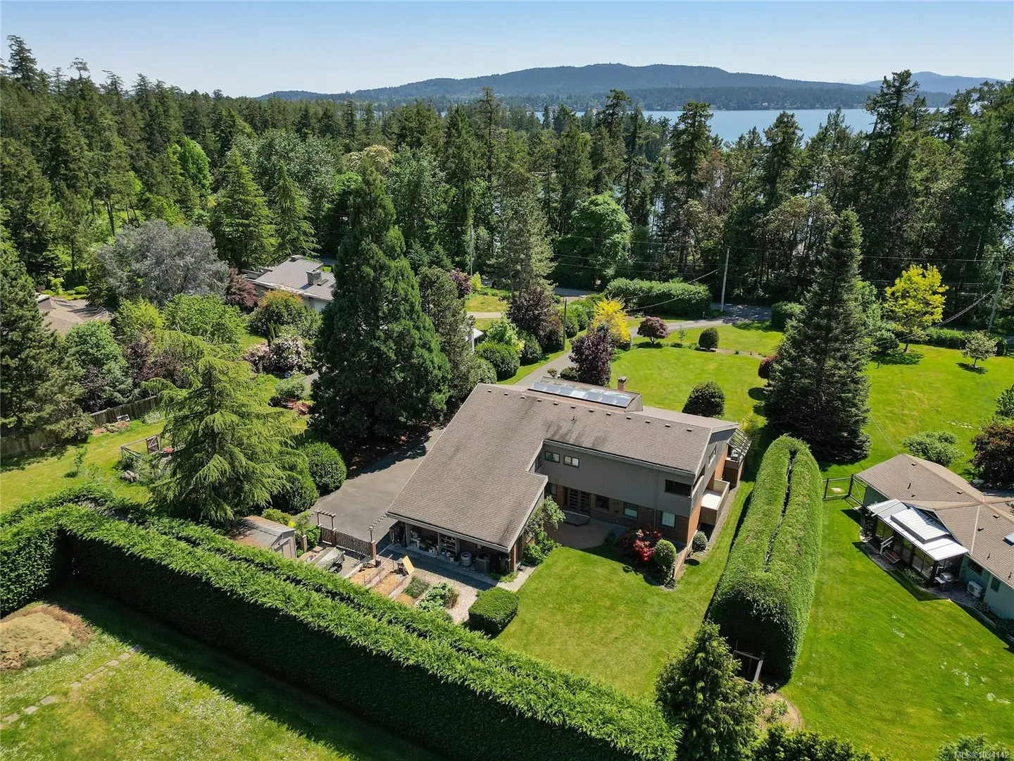 Aerial view of a gray house with a brown roof, surrounded by green trees and lawn, with a lake in the background.