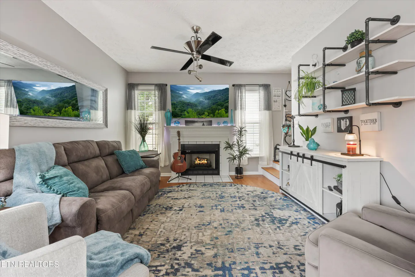 Living room with gray walls, a brown sofa with blue pillows, a fireplace, and a blue and gray rug. A guitar stands near the fireplace.