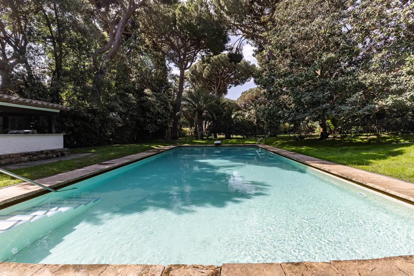 Rectangular pool with clear blue water, surrounded by green trees and grass. Steps lead into the pool. A white building is visible on the left.