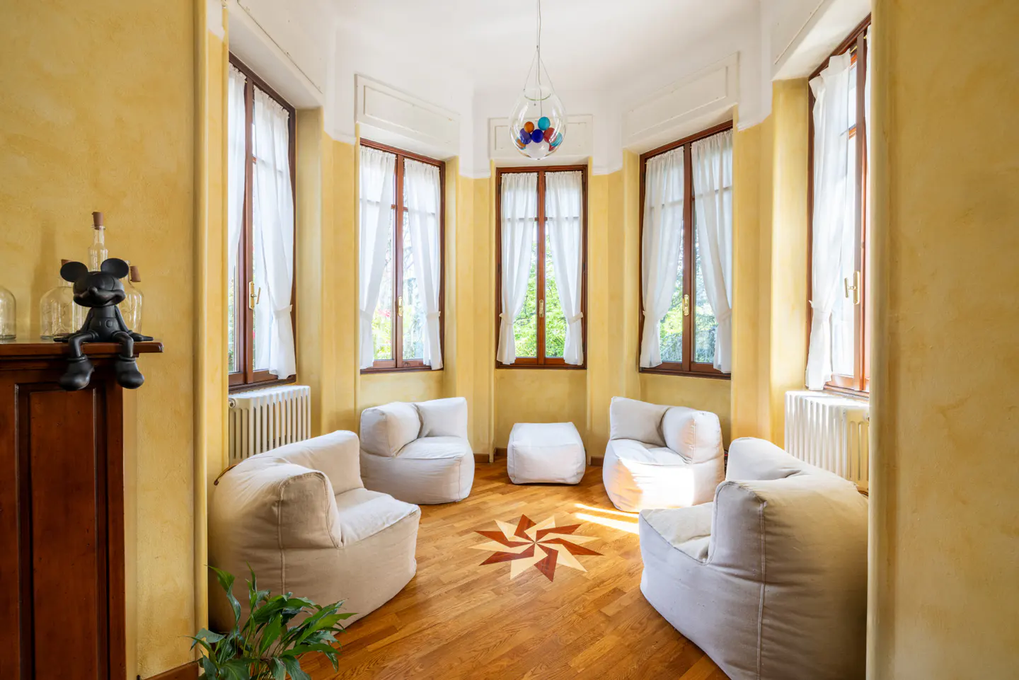 Sitting room with yellow walls, wood floors, and white chairs arranged around a starburst pattern. Windows with white curtains let in natural light.