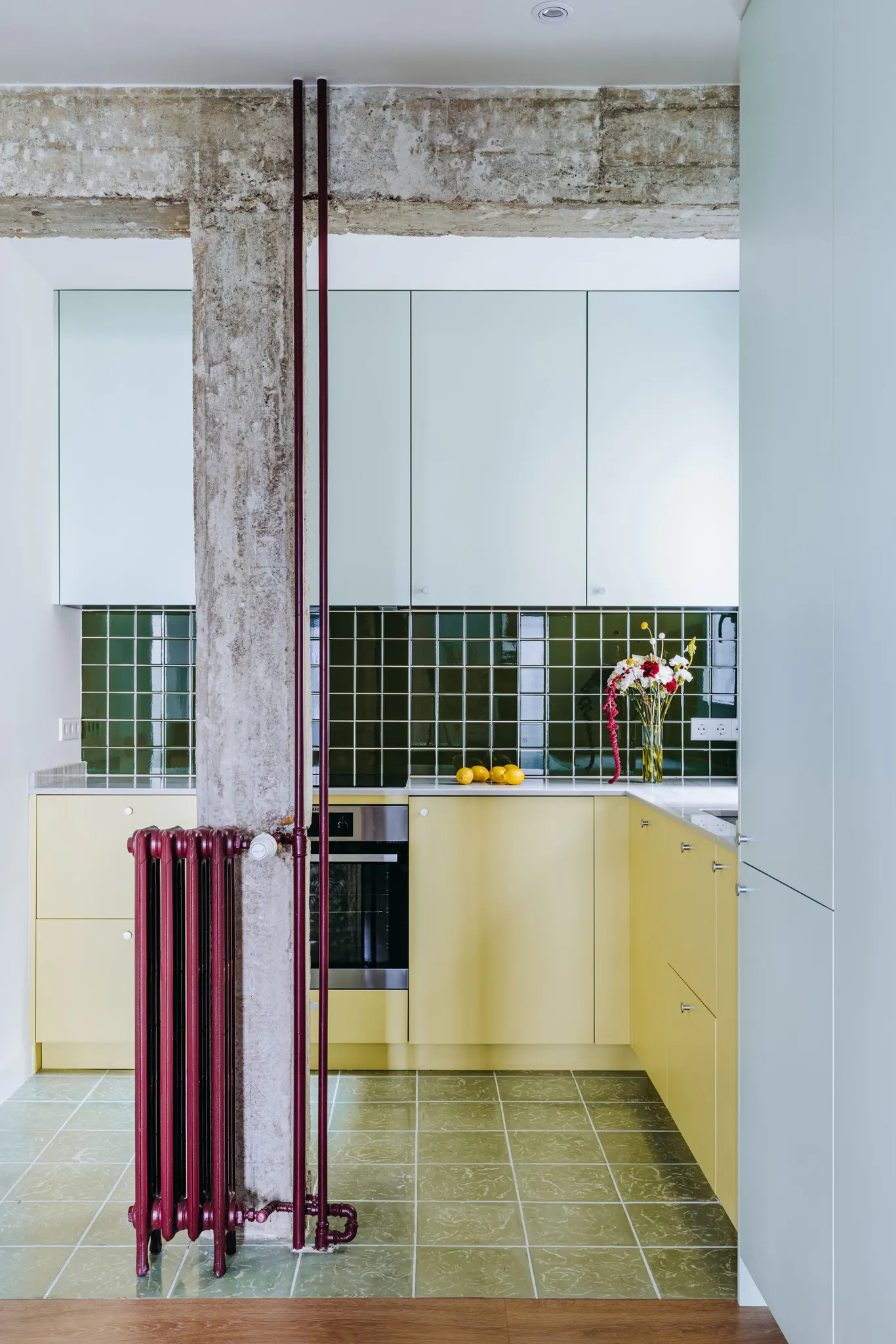 A modern kitchen with yellow cabinets, green tile backsplash, and a red radiator next to a concrete pillar.