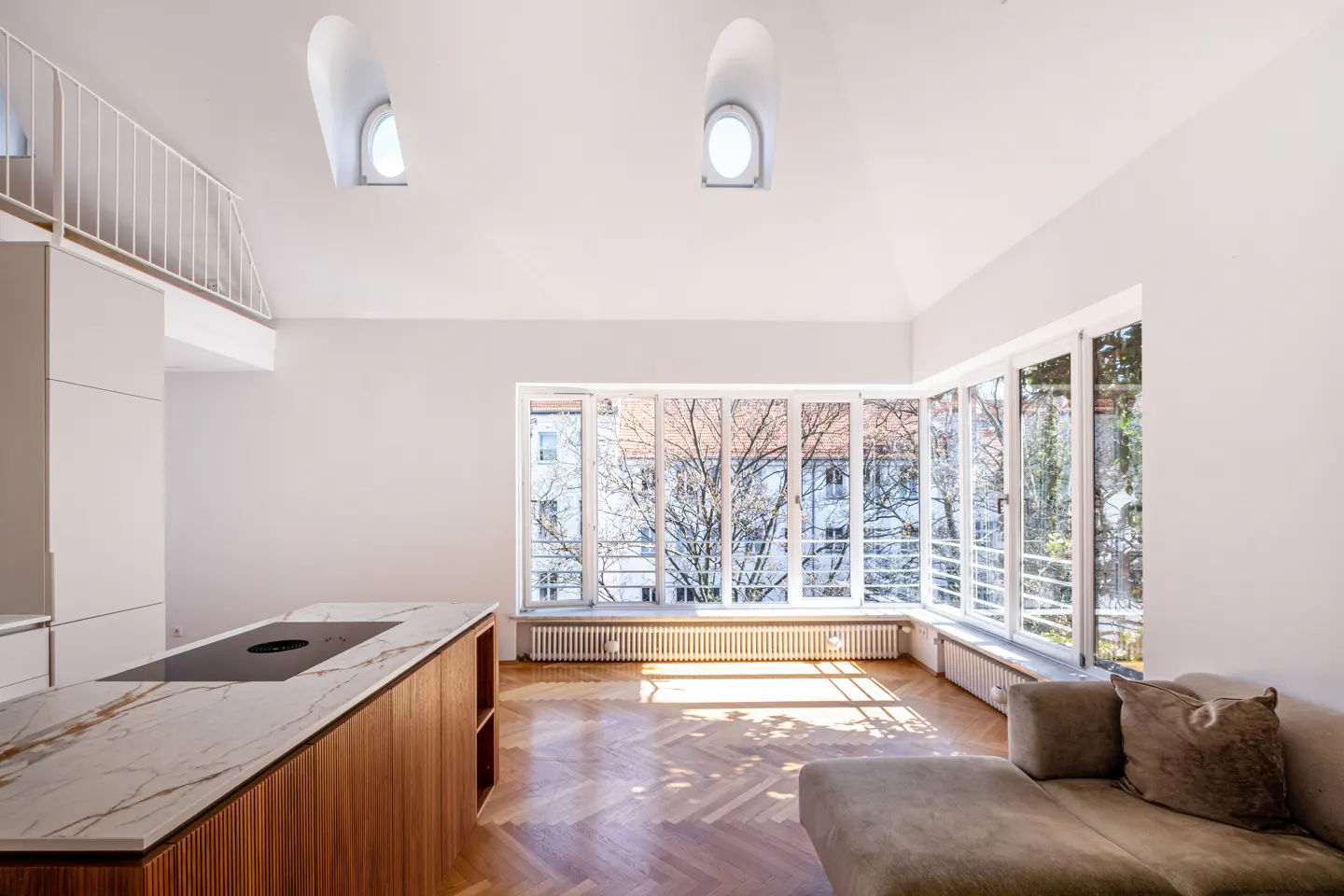 Bright, modern apartment interior with herringbone wood floors, white walls, and large windows. Kitchen island and gray sofa visible.