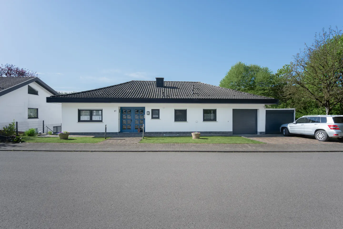 A white single-story house with a blue door, a black roof, and a gray garage door. A silver car is parked in the driveway.