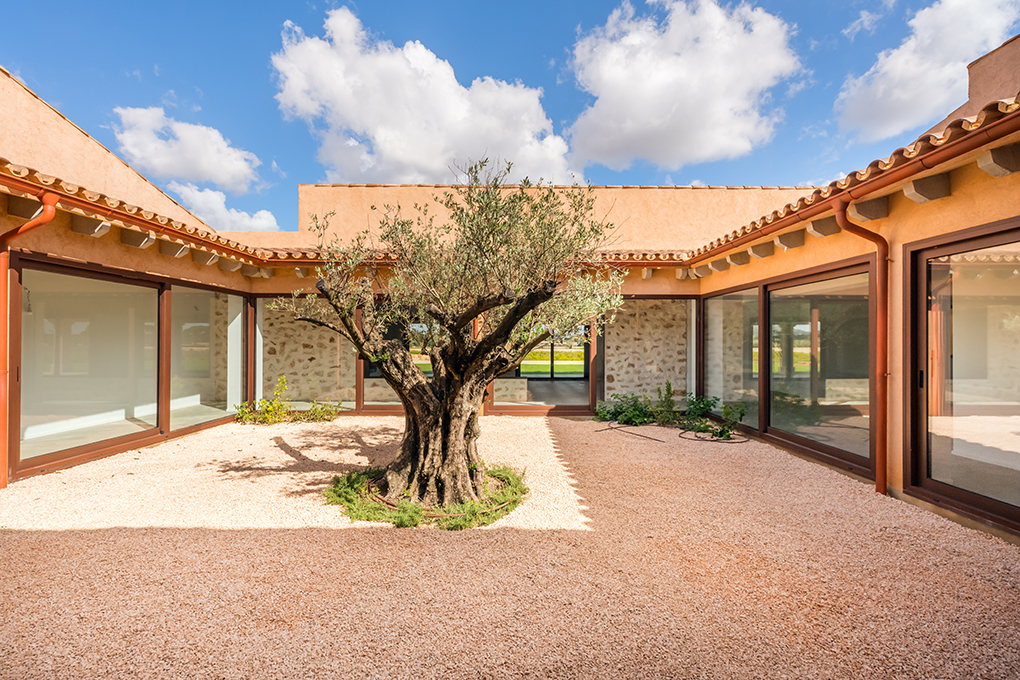 Courtyard view of a peach-colored house with a large olive tree in the center. The ground is covered in small peach-colored stones.