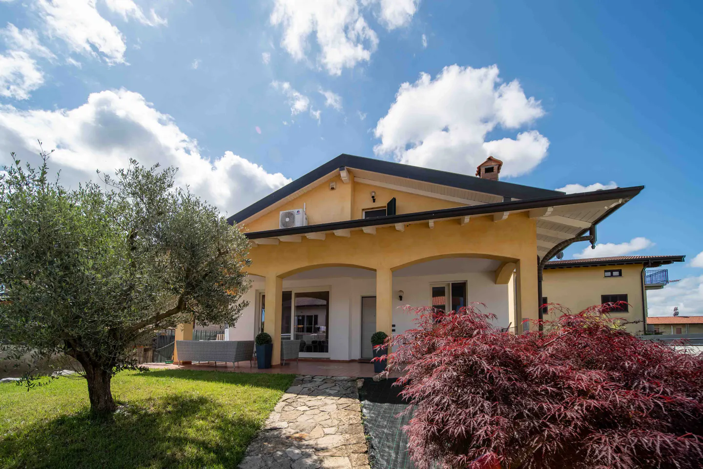 Two-story yellow house with a covered porch, stone walkway, and green lawn under a blue sky with white clouds.