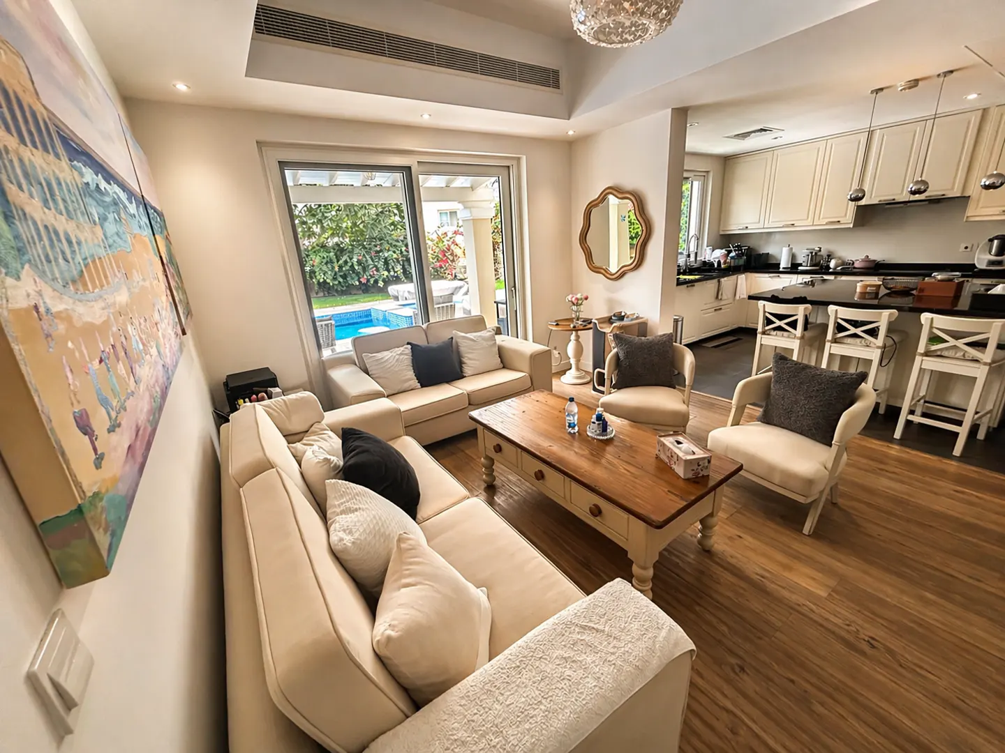 Living room with beige sofas, wooden coffee table, and chairs. Kitchen with white cabinets and bar stools in the background. Pool visible through sliding glass doors.