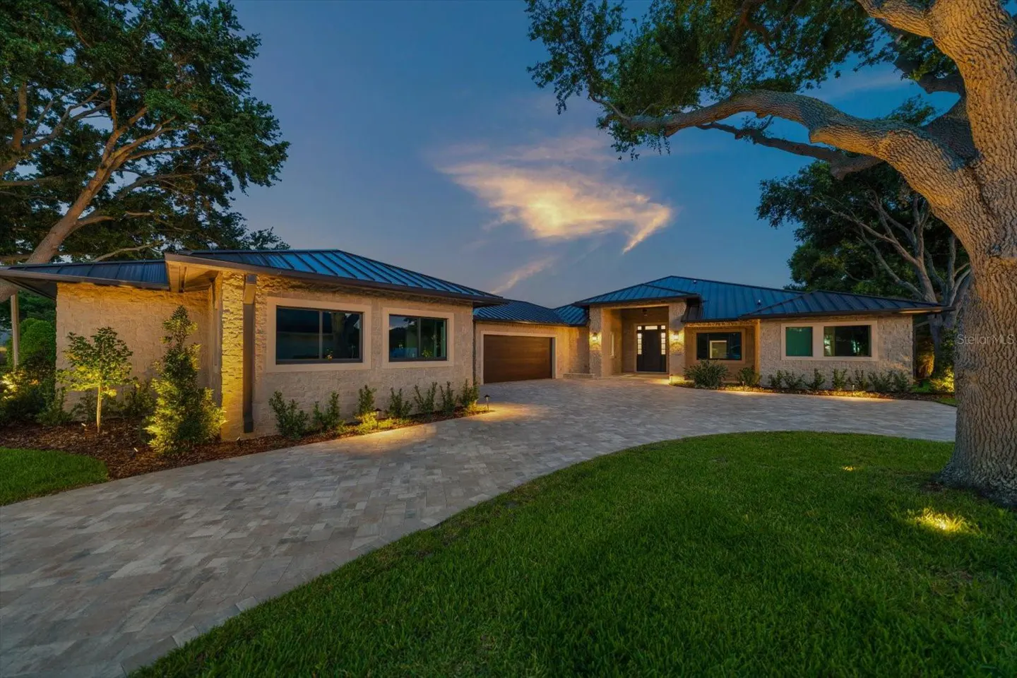 Exterior view of a tan, modern home with a blue roof, a stone driveway, and green grass at dusk.