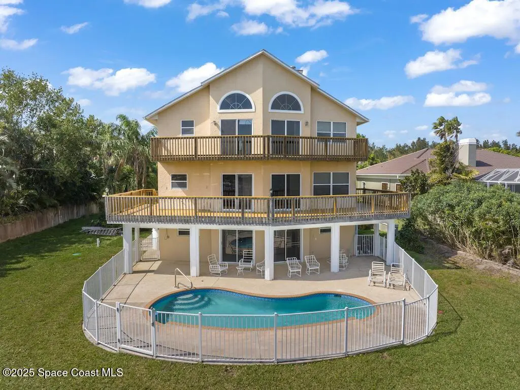Three-story tan house with two large wooden balconies, a fenced-in kidney-shaped pool, and patio furniture on the lawn.