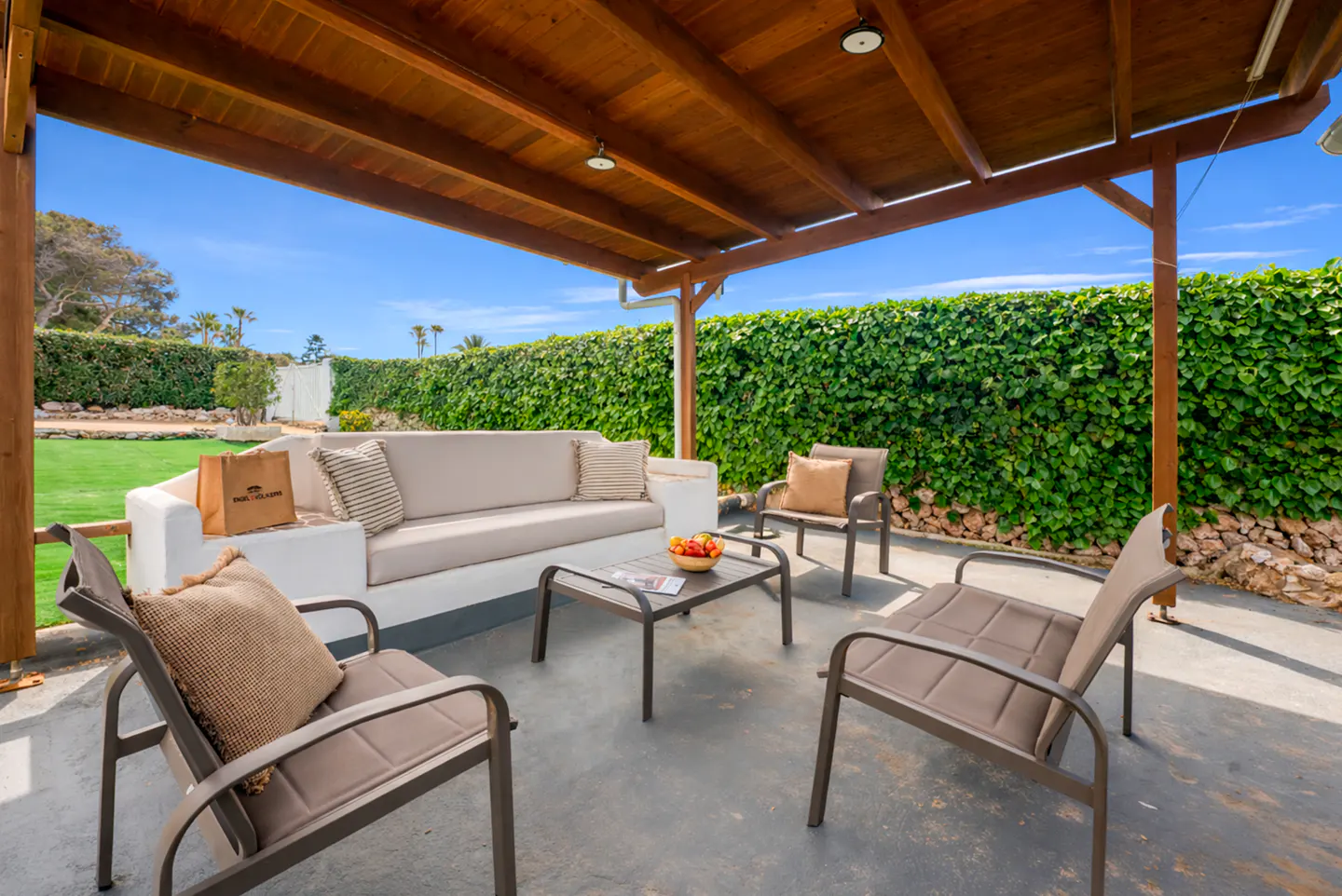Outdoor patio with a wooden roof, sofa, chairs, and a table with fruit. A green hedge is in the background.