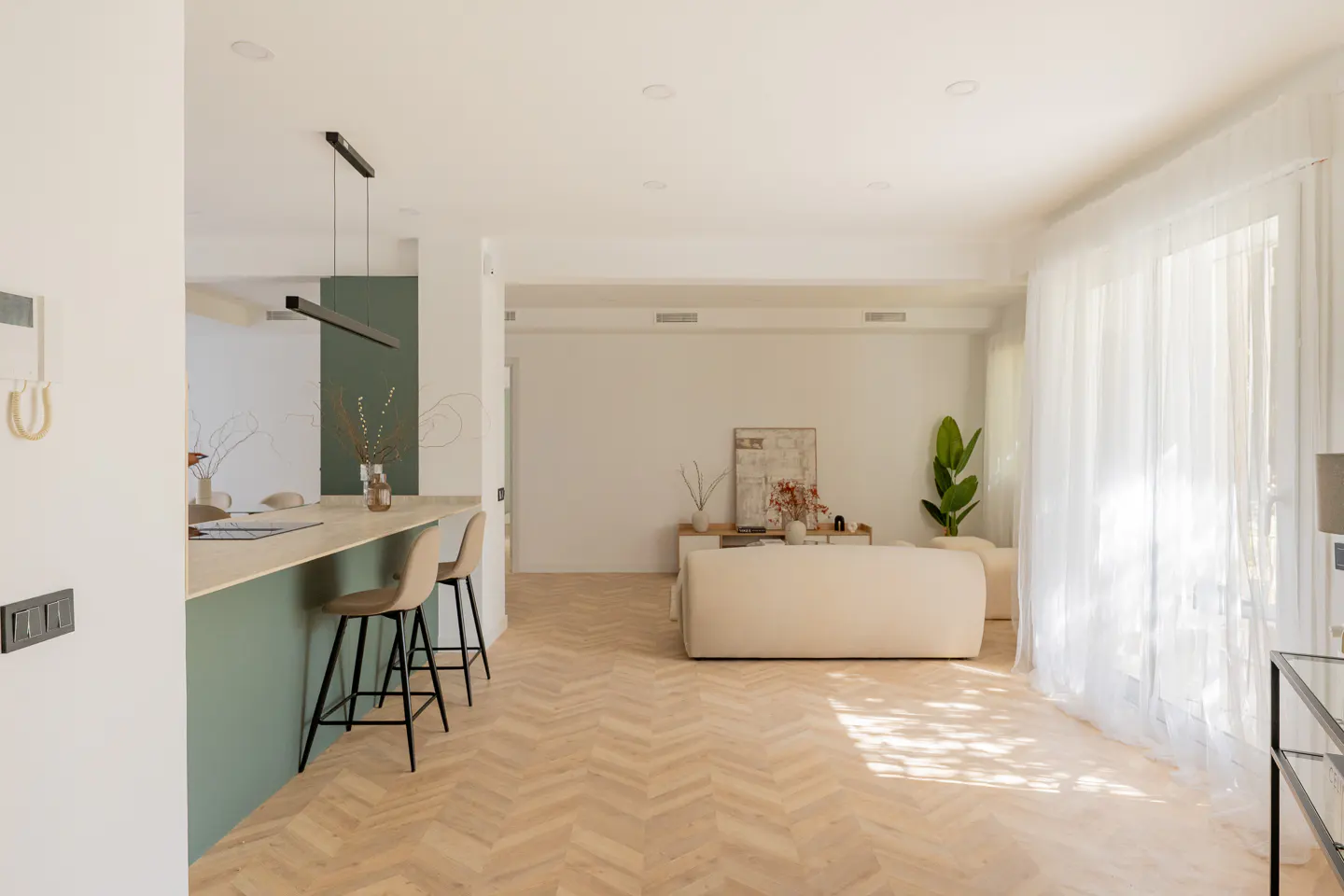 Bright, modern living space with herringbone wood floors, a cream sofa, and a green kitchen island with bar stools. Sunlight streams through sheer white curtains.