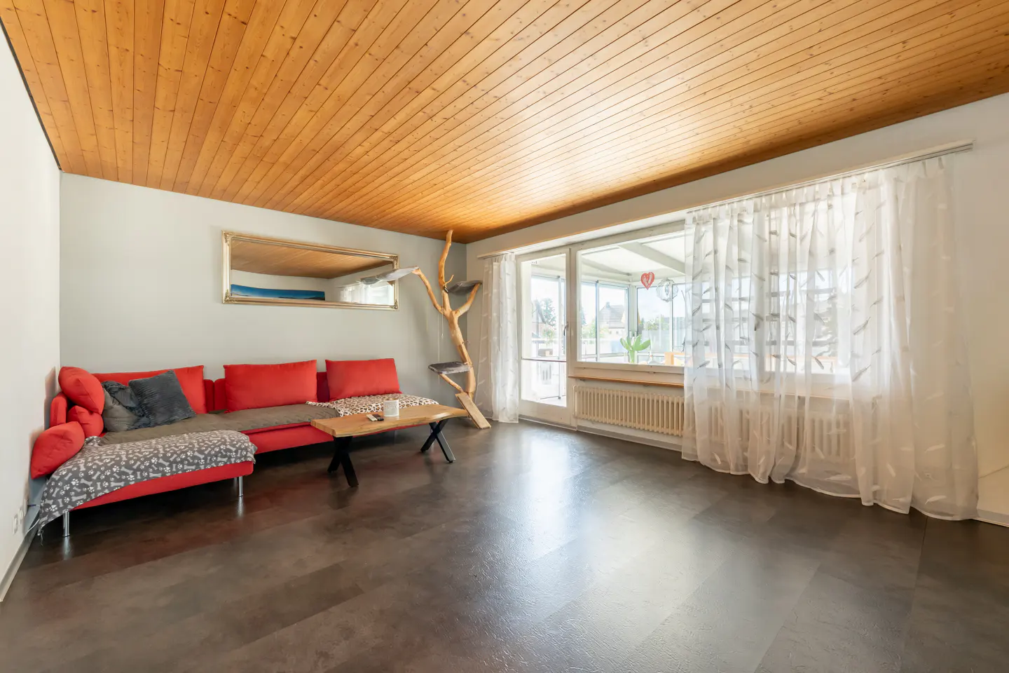 Living room with a red sectional sofa, wooden coffee table, cat tree, and large windows with white curtains under wood ceiling.