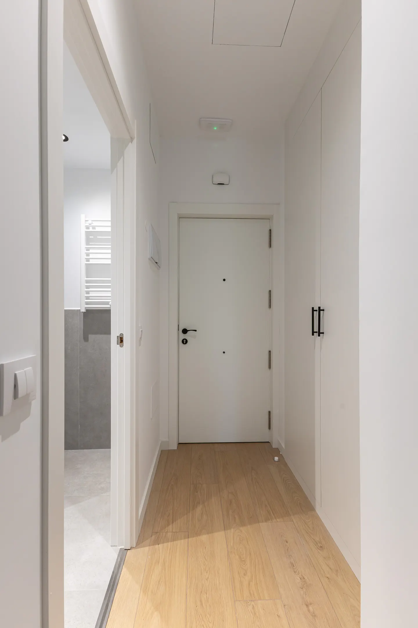 Hallway with light wood floors, white walls, and white doors. A bathroom is visible through an open doorway.