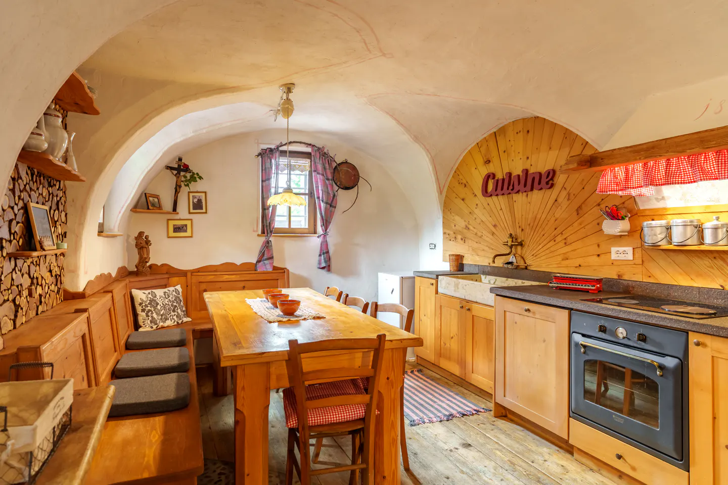 Rustic kitchen with vaulted ceiling, wood cabinets, and a long wooden table with bench seating. A "Cuisine" sign adorns a wood-paneled wall.