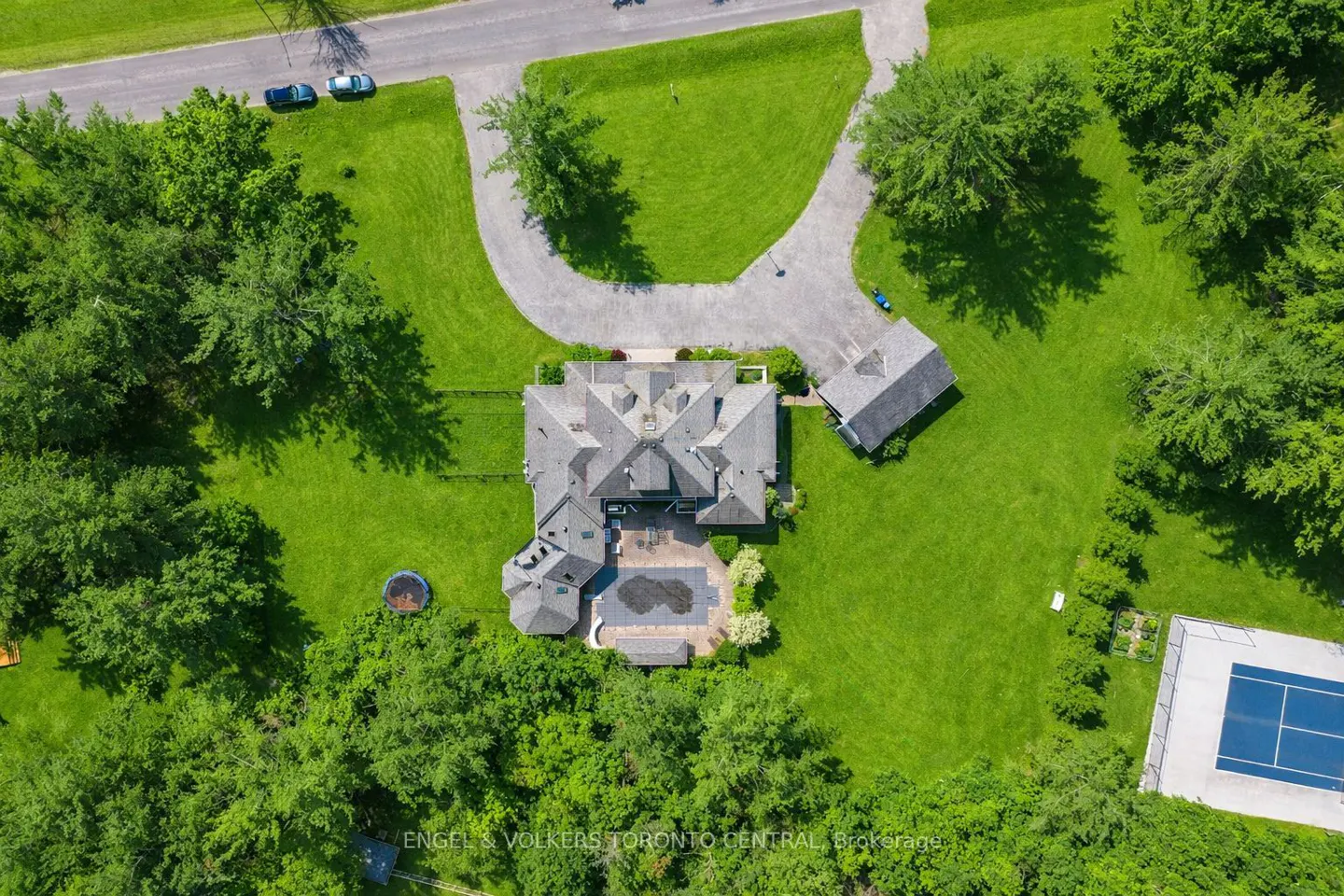 Aerial view of a large gray house with a pool, tennis court, and long driveway surrounded by green grass and trees.