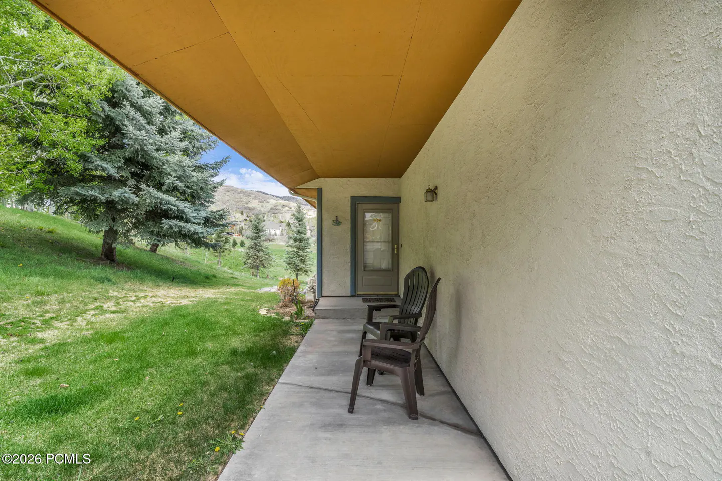 Covered porch with two dark chairs, a door, and a view of a green lawn and trees.