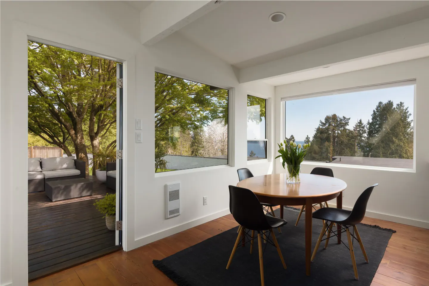 Bright dining room with wood floors, black rug, round table, and four black chairs. Large windows overlook trees and a patio with outdoor seating.