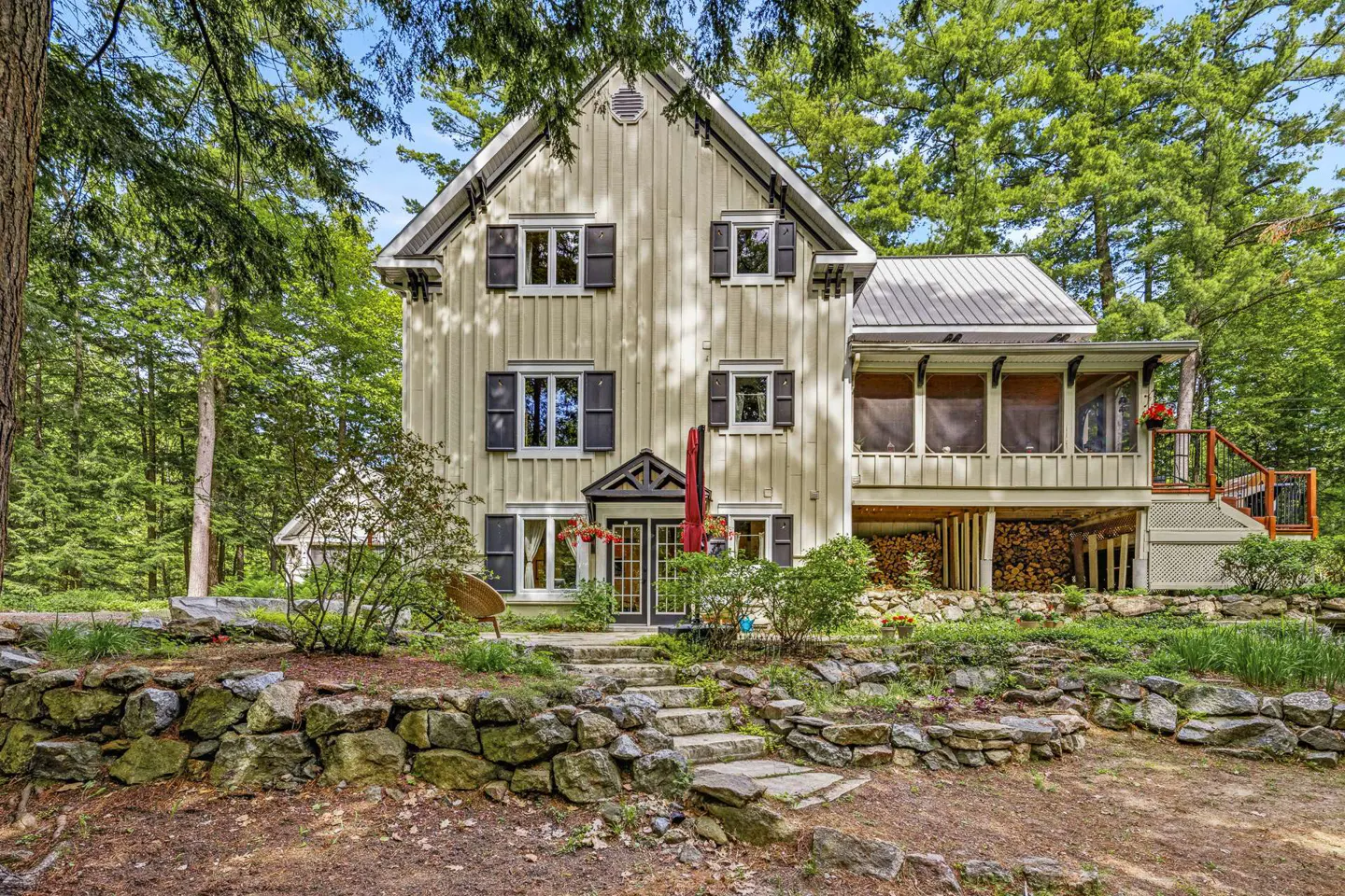 Three-story cream-colored house with dark shutters, a screened porch, and a stone retaining wall in a wooded setting.