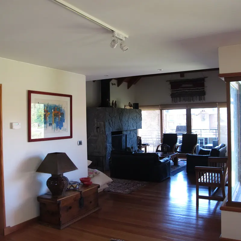 Living room with hardwood floors, stone fireplace, and large windows. A painting hangs above a wooden chest with a lamp. Sofas and chairs provide seating.