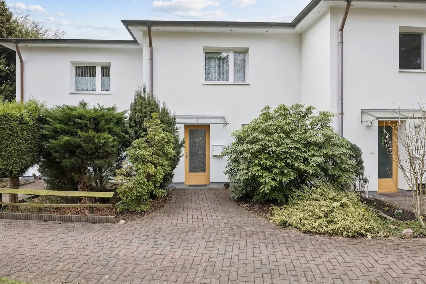 Exterior view of a modern, white townhouse with a brick driveway and green landscaping.
