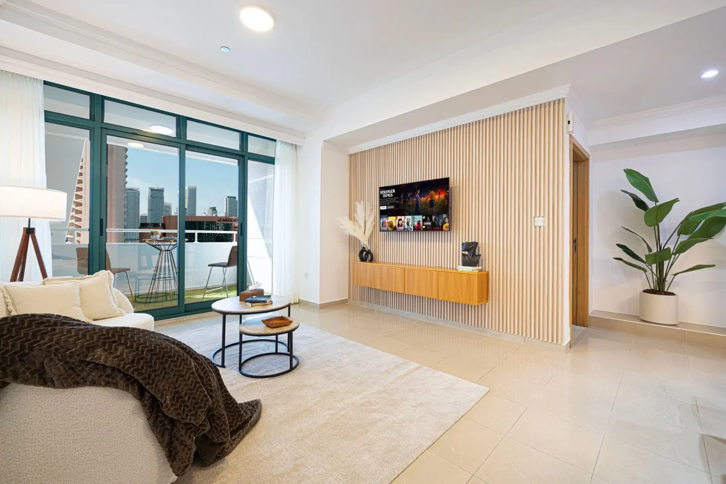 Bright living room with a white sofa, brown throw, and a balcony overlooking city buildings. A TV is mounted above a wooden cabinet.