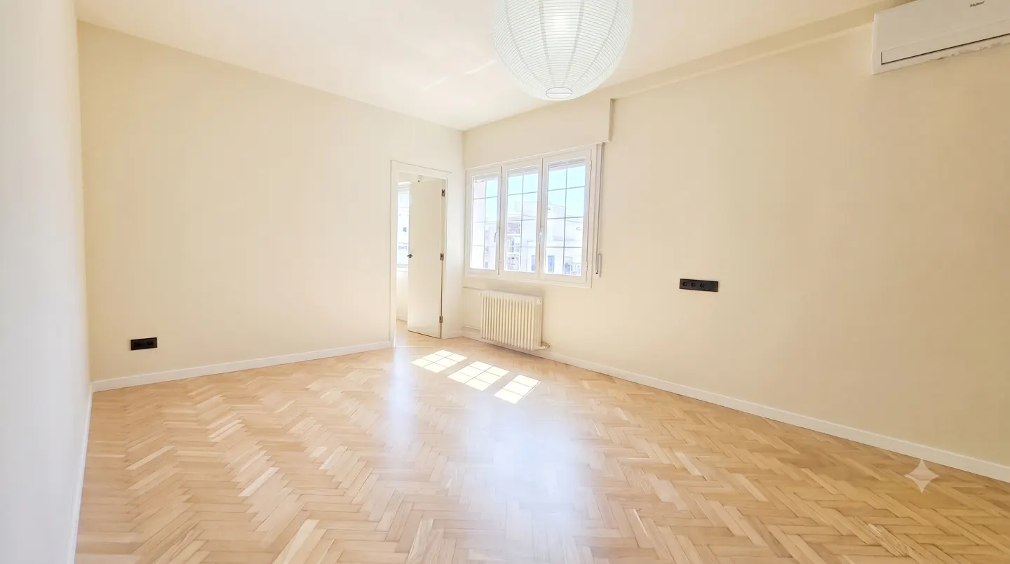 Empty room with light beige walls, herringbone wood floors, a white paper pendant lamp, a window, and an AC unit.