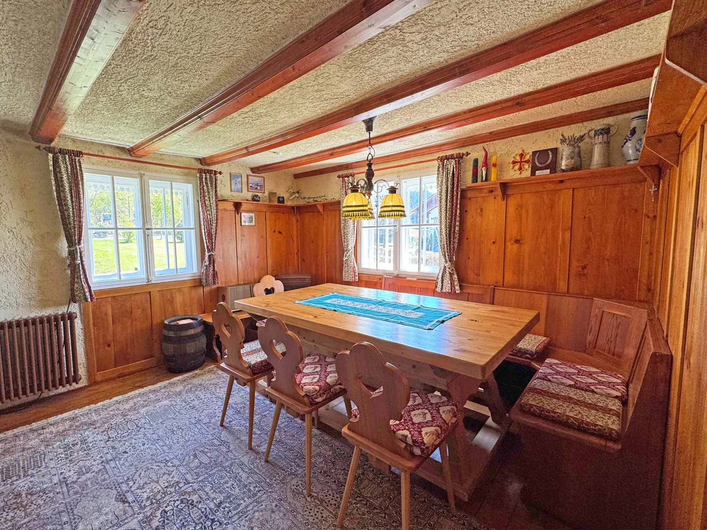 A rustic dining room with wood paneling, beams, and a large wooden table with chairs and a bench. Two windows let in natural light.