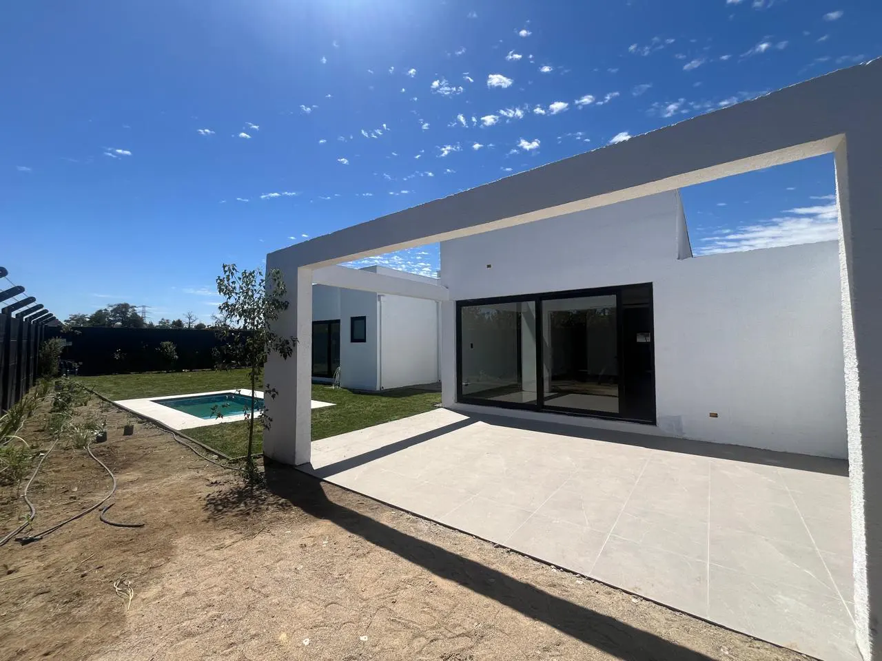 Modern white house with a patio and pool under a blue sky. Large sliding glass doors open to the patio. A small tree is near the pool.