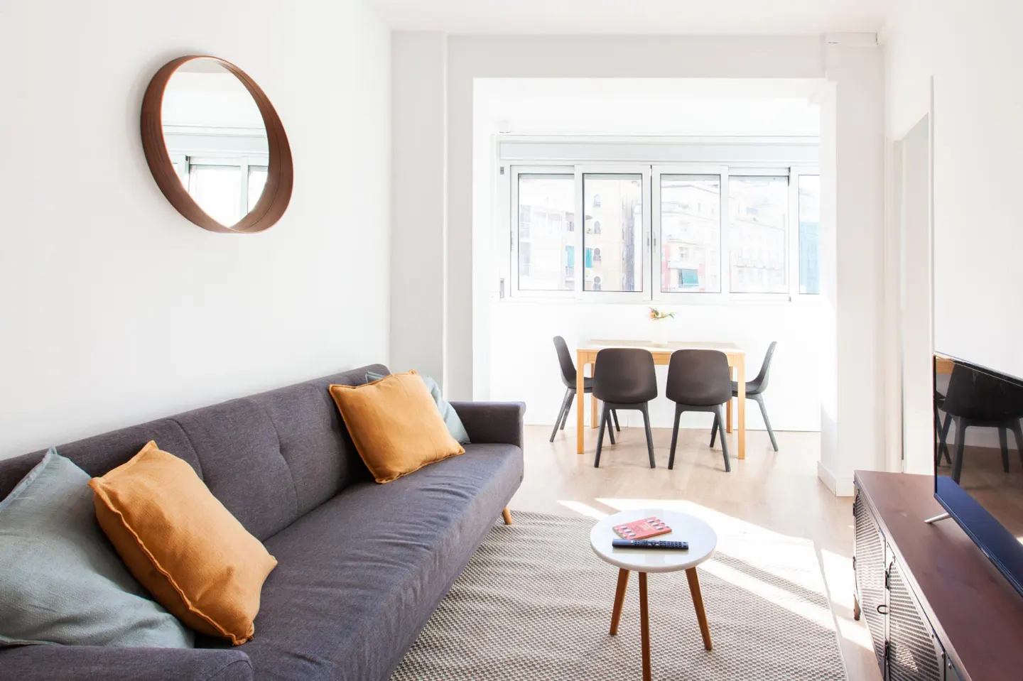 Bright living room with gray sofa, orange pillows, round mirror, and dining table near a window.