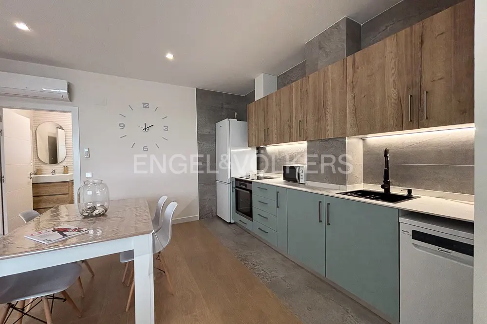 A modern kitchen with light blue cabinets, wood upper cabinets, and a white refrigerator. A dining table with white chairs is in the foreground.