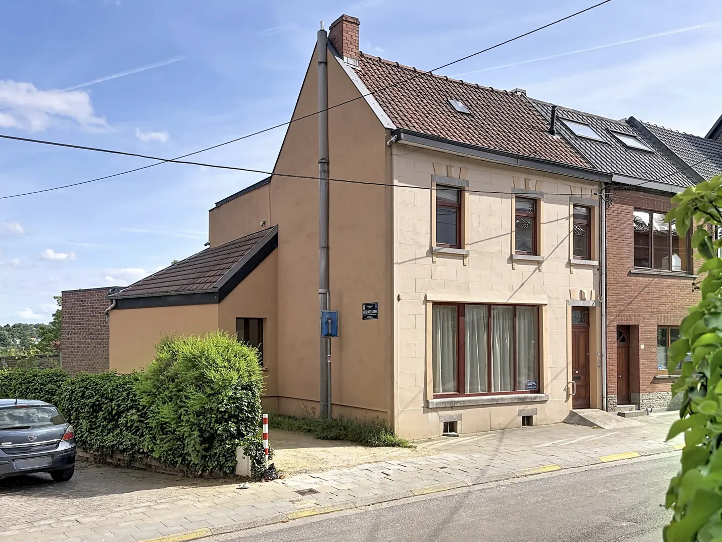 A two-story beige house with a brown tiled roof, large windows, and a small side extension next to a street.