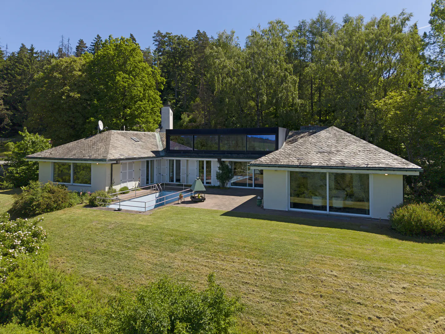 A modern white house with a pool and a large lawn, surrounded by green trees.