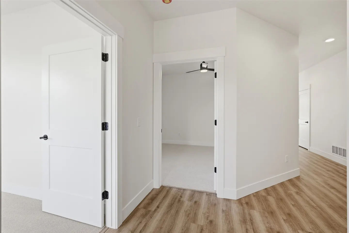 Bright hallway with wood floors and white walls. Two open white doors lead to carpeted rooms. A ceiling fan is visible in one room.