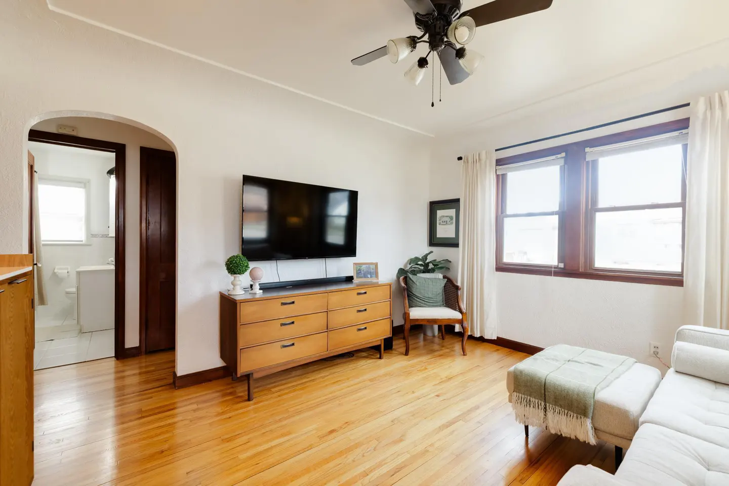Living room with hardwood floors, white walls, and a TV mounted above a wooden dresser. A doorway leads to a bathroom. A ceiling fan is visible.